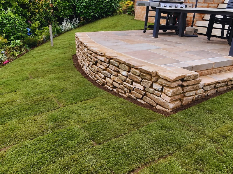 A raised paved patio with a dry-stone-effect retaining wall, built as an outdoor dining and cooking space off the lawn