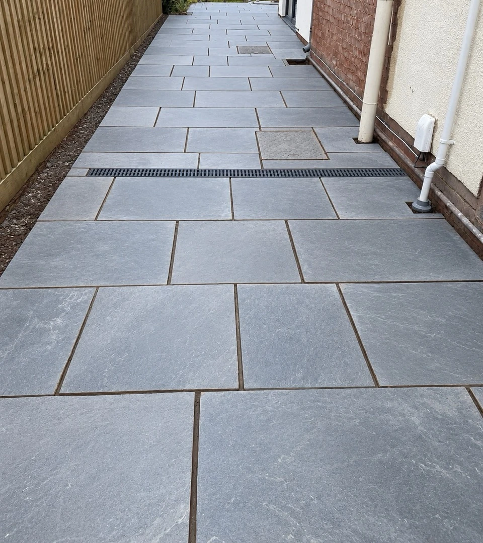 A stone path with a discreet drainage channel running down the side of a house, opening onto a view over the fields beyond