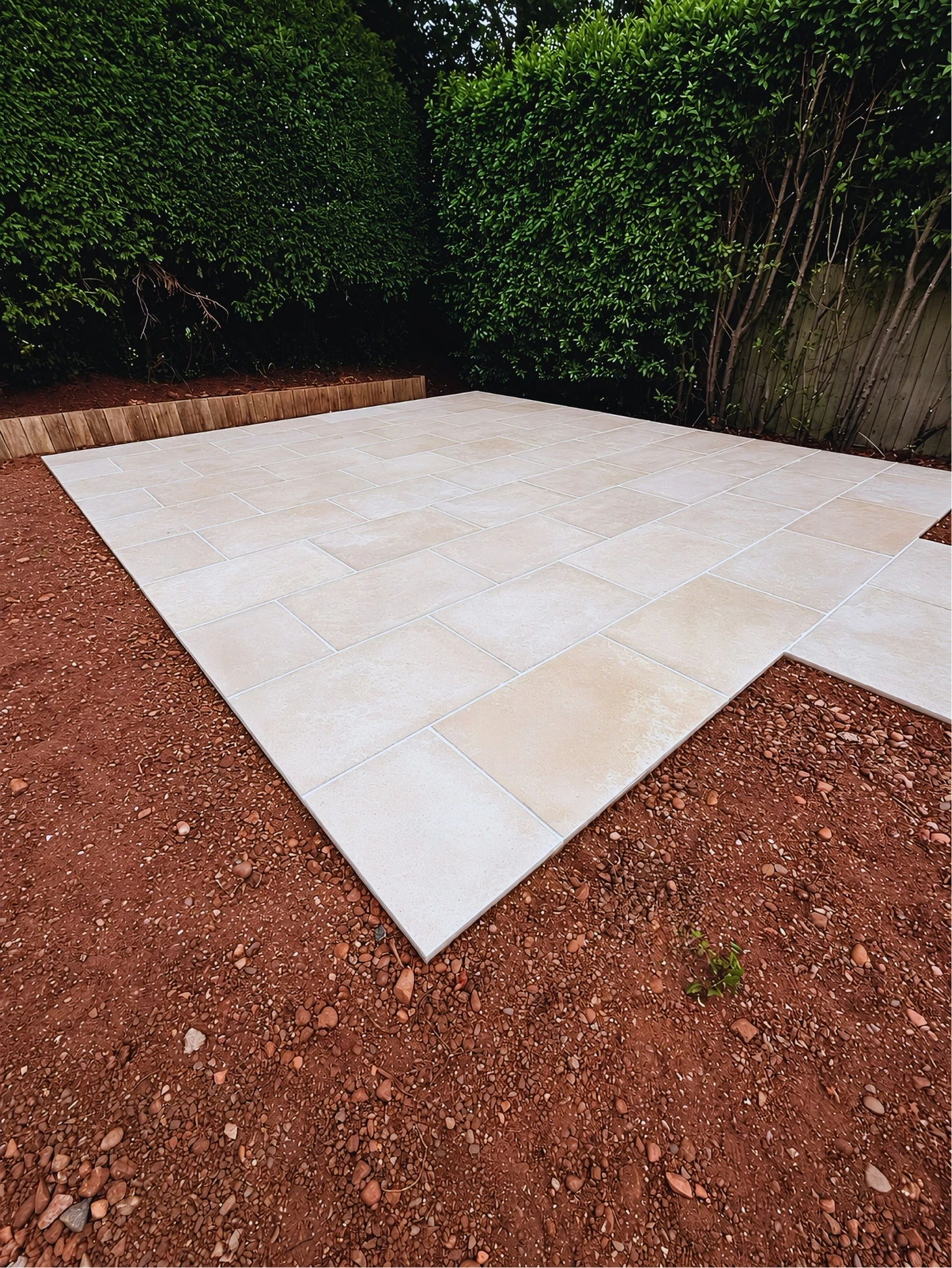 The paved base for the pergola, with a timber edge along the hedge behind it and bare soil on every other side.