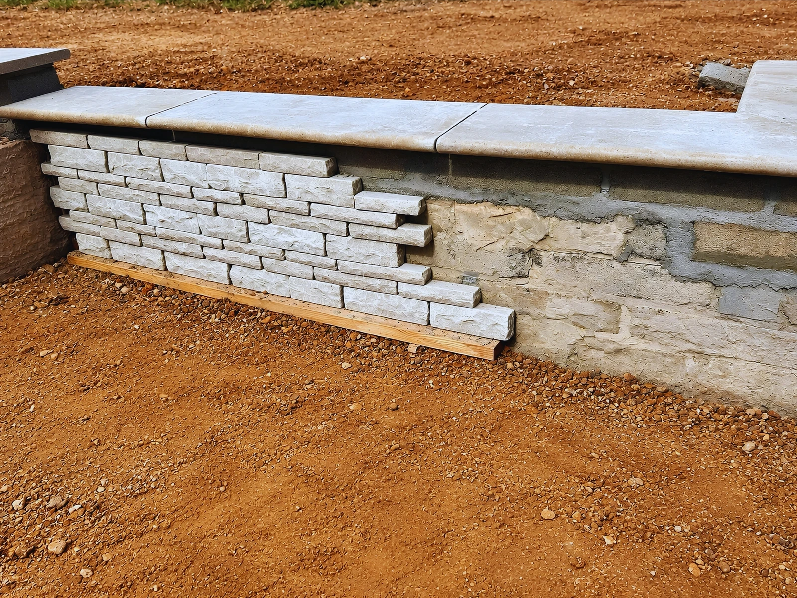 A retaining wall being built up in stacked stone, with drainage and backfill visible behind the coping already laid
