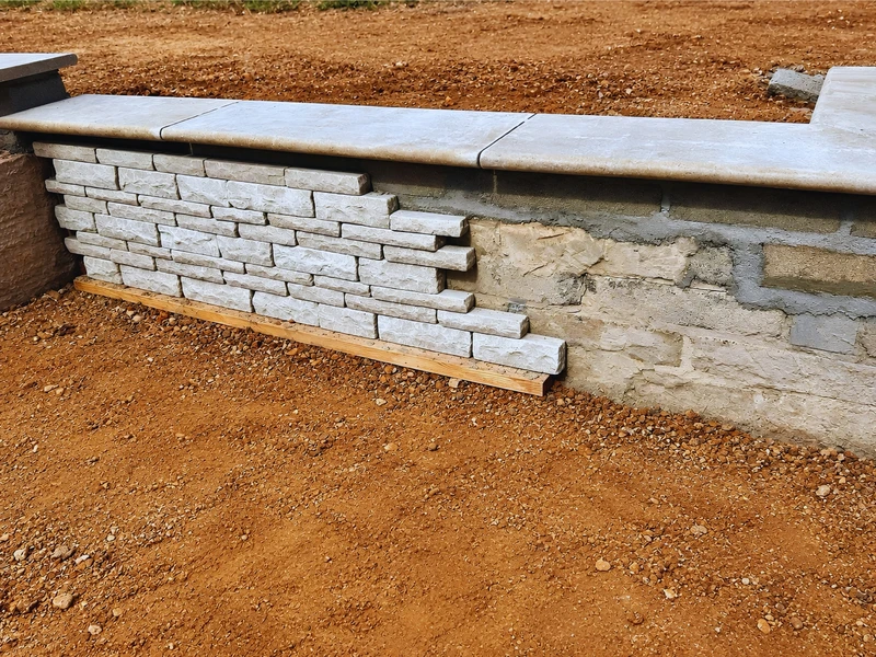 A retaining wall being built up in stacked stone, with drainage and backfill visible behind the coping already laid