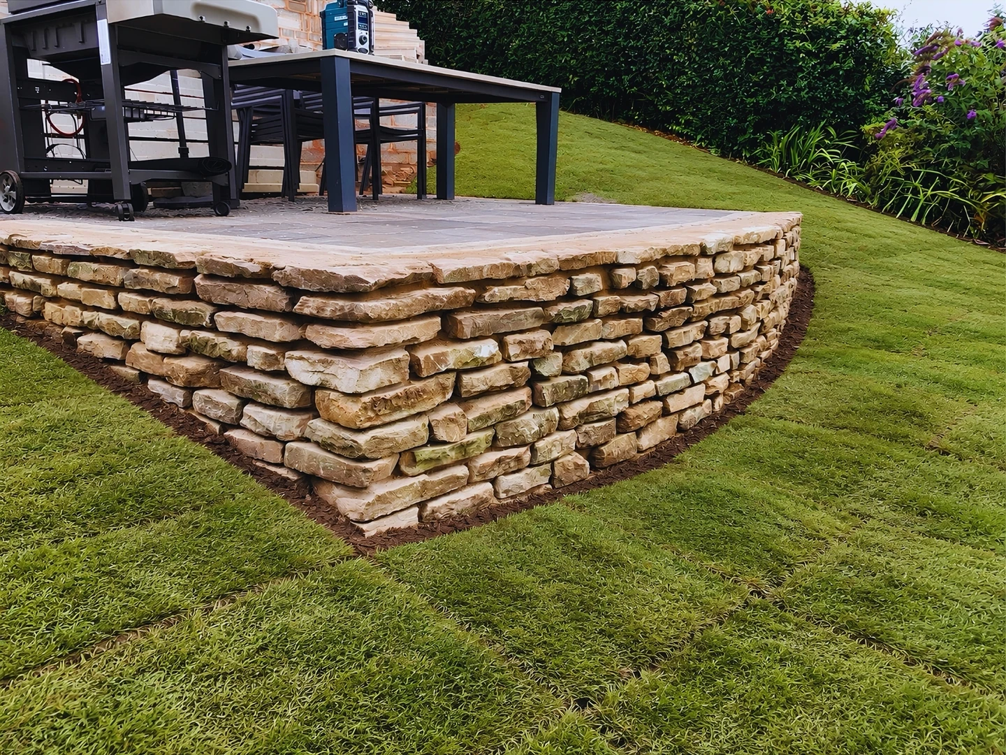 A raised paved seating area edged with a dry-stone-effect retaining wall, with newly turfed lawn around it
